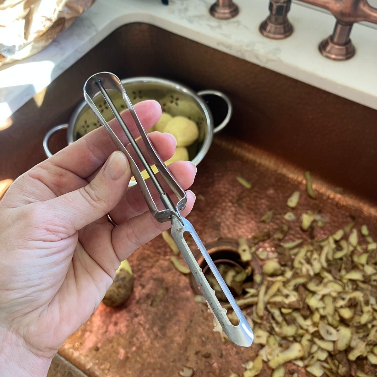 A hand holding a stainless steel peeler over a kitchen sink.