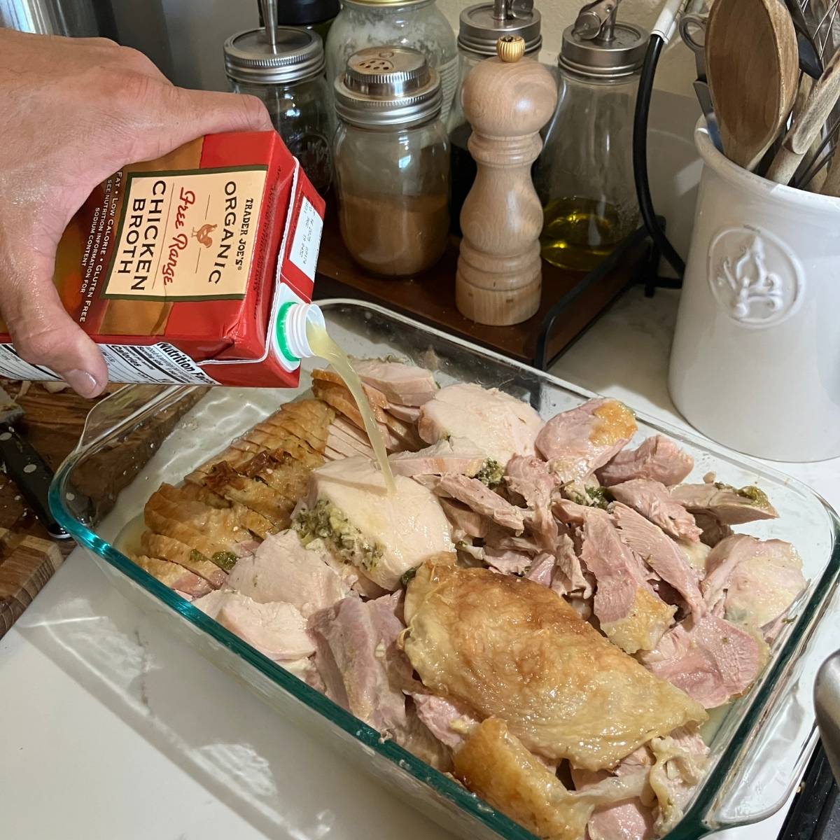 A mans hand pouring a box of chicken stock over a glass pyrex dish filled with roasted carved turkey.